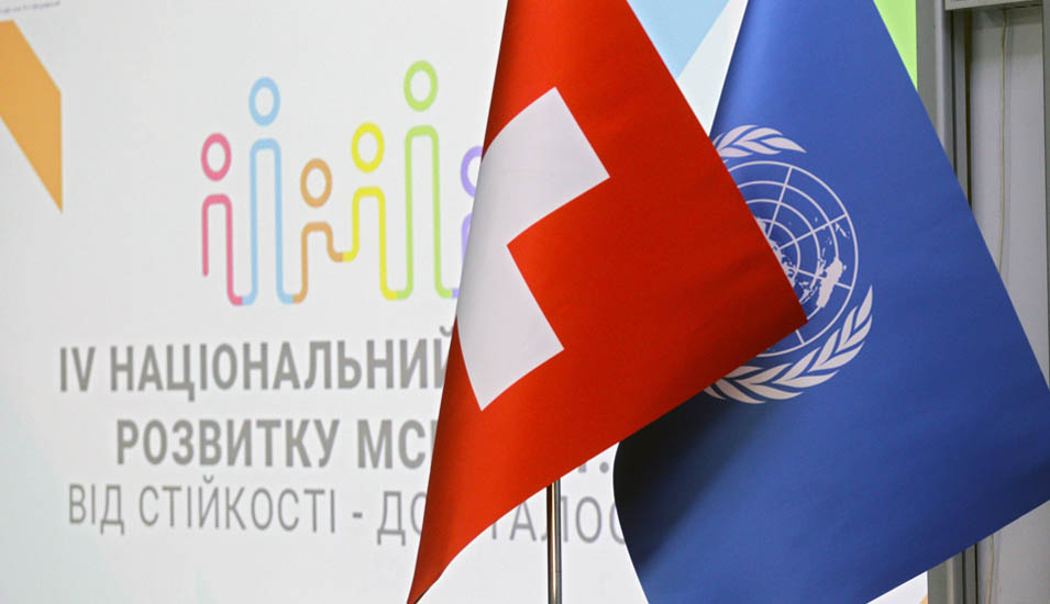 Aufnahme der Flaggen der Schweiz und der Vereinten Nationen vor einer Tafel mit ukrainischer Schrift.