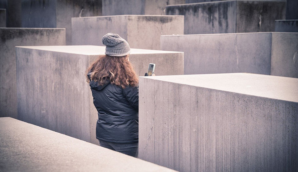 Eine junge Frau ist mit Mantel und Mütze von hinten zu sehen zwischen vielen grauen Steinquadern. Sie fotografiert mit ihrem Smartphone. 